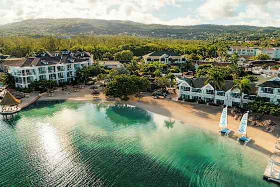 Flera hotellbyggnader vid en lugn sandstrand med klart vatten och utsikt över grönskande kullar. (AI-genererad text)