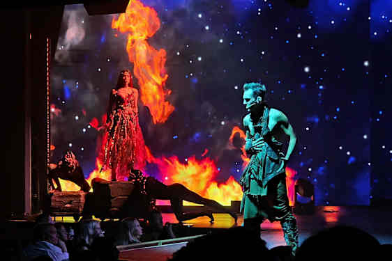 En scen med en kvinna i en eldklädsel omgiven av lågor och en man i en svart dräkt, med en stjärnbeströdd bakgrund. (AI-genererad text)