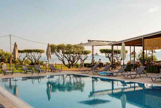 Utsikt över en pool med solstolar runtom, träd i bakgrunden och havet i fjärran. (AI-genererad text)