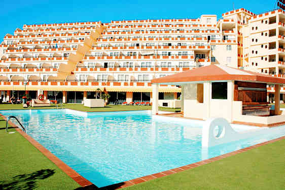 En stor pool omgiven av grönområden och ett hotell med flera våningar i bakgrunden. (AI-genererad text)