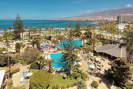 Utsikt över ett semesteranläggning med pooler, palmer och människor som solar och badar nära havet. (AI-genererad text)