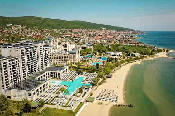 Hotell och pooler vid stranden med havet och staden i bakgrunden. (AI-genererad text)