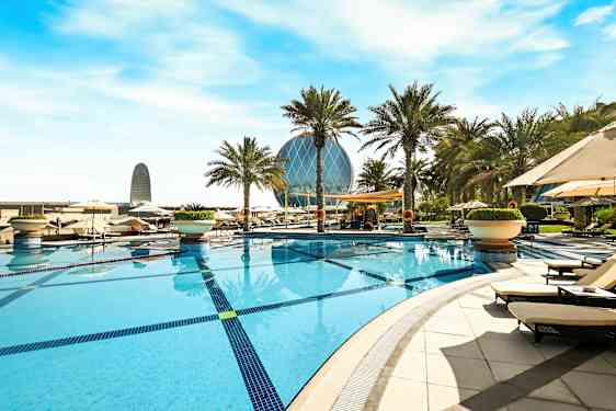 En swimmingpool omgiven av solstolar och palmer med en modern byggnad i bakgrunden. (AI-genererad text)