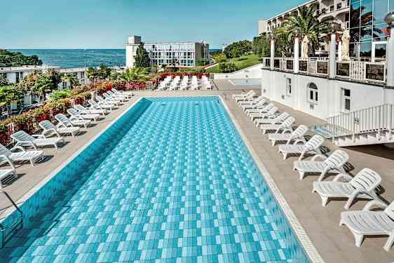 En rektangulär pool omgiven av solstolar, med havet och en byggnad i bakgrunden. (AI-genererad text)