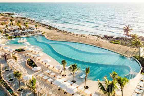 En stor pool omgiven av solstolar och parasoller ligger vid stranden med utsikt över havet. (AI-genererad text)