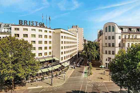 Ett stadslandskap som visar en byggnad med texten "Bristol" på taket, omgiven av träd och en gata med uteservering. (AI-genererad text)