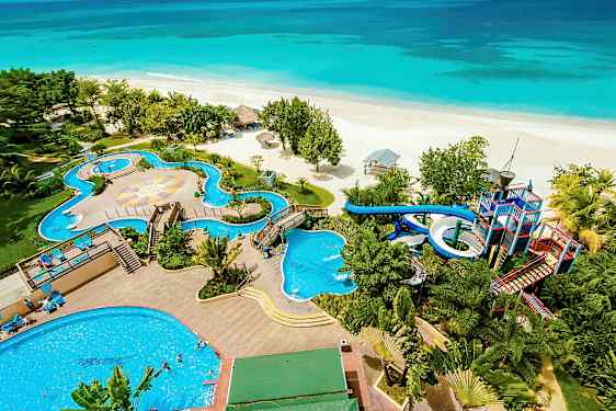 Bilden visar en strand framför en resort med pooler, vattenrutschbanor och omgivande grönska. (AI-genererad text)