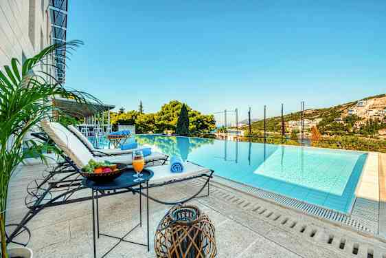 En utsikt mot en infinitypool med solstolar, bord med frukt och dryck intill sig, samt gröna omgivningar och himmel i bakgrunden. (AI-genererad text)