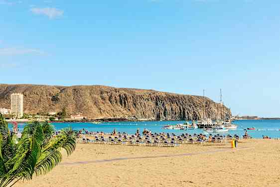 Strand med solstolar, havet och båtar vid en klippa. (AI-genererad text)