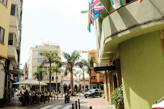 En gata med byggnader, palmer och en hotellskylt. Människor sitter vid ett kafé och ett motorfordon kör förbi. (AI-genererad text)