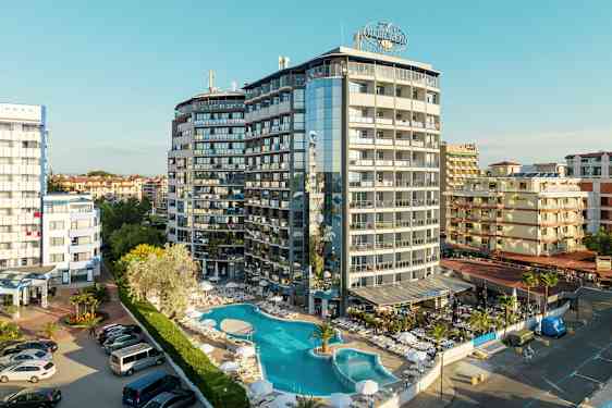 Ett flervåningshotell med pool och solstolar framför, inbäddat i ett tätbebyggt område. (AI-genererad text)