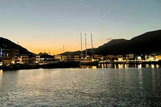 En hamn i skymningen med båtar förtöjda vid en kaj och upplysta byggnader på avstånd. Berg syns i bakgrunden. (AI-genererad text)