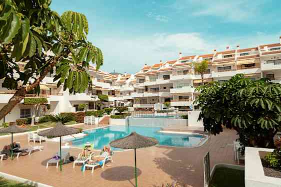 Ett utomhusområde med en pool, solstolar och ett flervåningshotell. Flera parasoller och gröna växter omger området. (AI-genererad text)