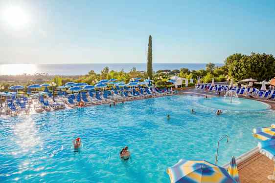 En swimmingpool med solsängar och parasoller omkring, omgiven av grönska och med havsutsikt. (AI-genererad text)