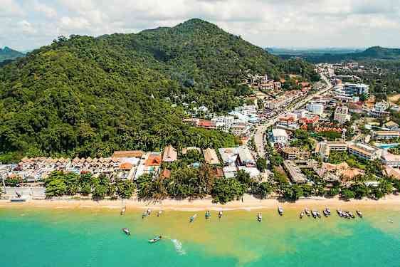 Flygfoto över en strand med båtar i vattnet och en stad mot grönskande kullar i bakgrunden. (AI-genererad text)