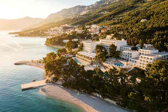 Luftfoto av hotell vid kusten och strand, omgiven av träd och med berg i bakgrunden. (AI-genererad text)