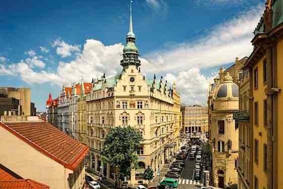 Ett europeiskt stadsmotiv med en dekorativ byggnad med torn, omgiven av fler historiska byggnader. Himlen är blå med vita moln. (AI-genererad text)