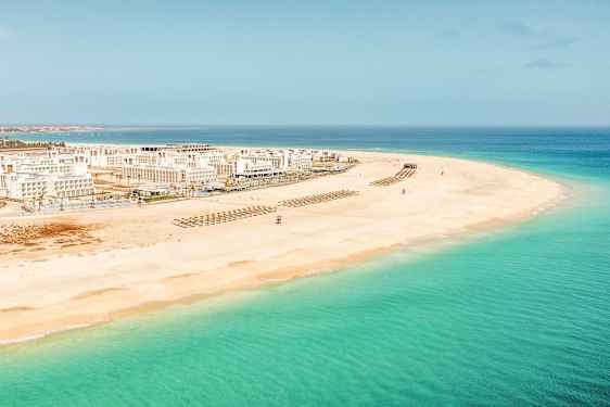 Flygfoto av en vit sandstrand med klart turkost vatten och vita byggnader i bakgrunden. (AI-genererad text)