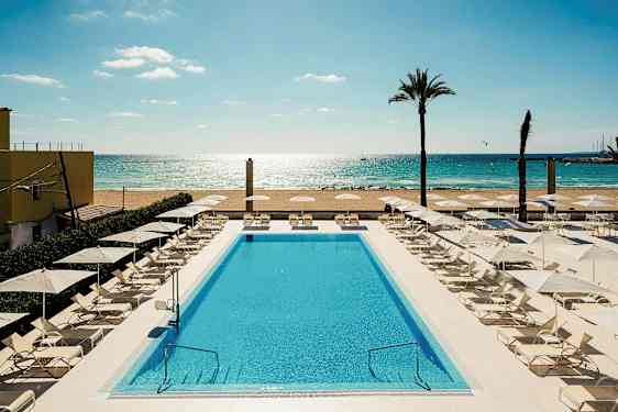 En pool omgiven av solstolar och parasoller nära en sandstrand och hav. (AI-genererad text)