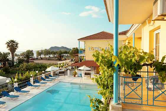 Bild av hotellpool med solstolar, byggnad med gul fasad och trädgård samt havet i bakgrunden. (AI-genererad text)