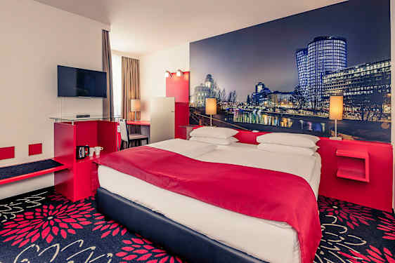 Ett hotellrum med en dubbelsäng, en TV på väggen och ett skrivbord bredvid fönstret. Väggarna är dekorerade med en storbild av en stadslandskap. (AI-genererad text)