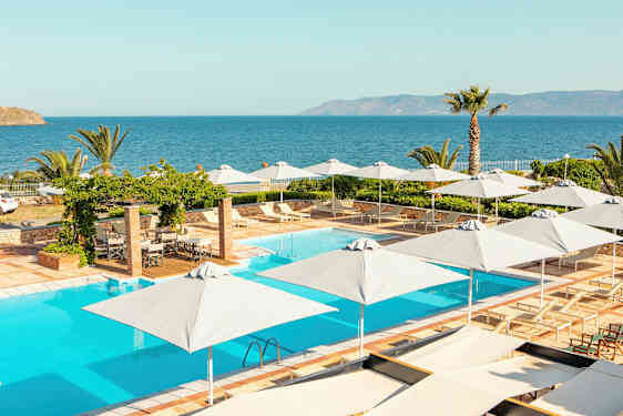 En pool med solstolar och parasoller, utsikt mot havet med växtlighet runt omkring. (AI-genererad text)