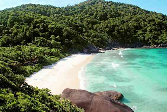 Bild på en strand omgiven av gröna berg och blått hav. (AI-genererad text)
