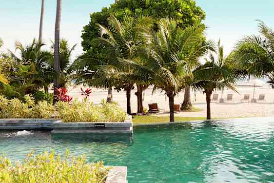 Poolområde med utsikt över en sandstrand och omgivande palmer. (AI-genererad text)