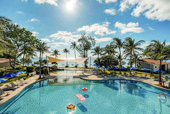 En pool omgiven av solstolar och parasoller, med utsikt över havet och palmer under en klar himmel. (AI-genererad text)