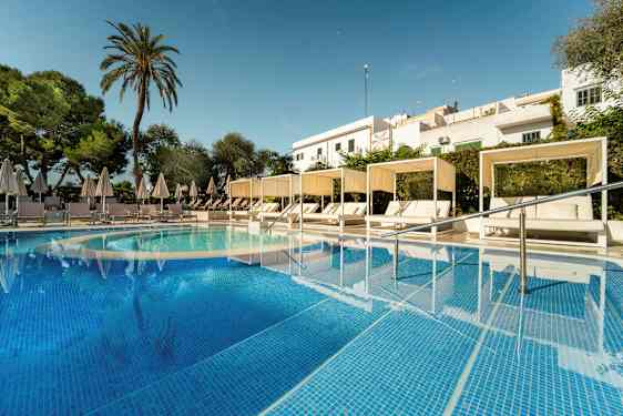 Bild av en pool med turkost vatten och vita solsängar under parasoll samt cabanas. Det är omgivande grönska och en skymt av byggnader i bakgrunden. (AI-genererad text)