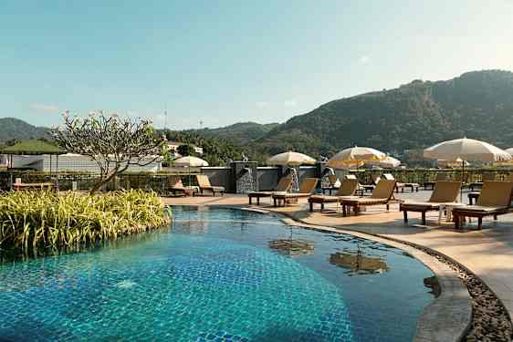 Utomhuspool med solstolar och parasoller, omgiven av grönska och berg i bakgrunden. (AI-genererad text)