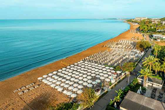 Bilden visar en sandstrand vid ett hav, med många parasoller och solstolar uppställda i en ordnad formation. I bakgrunden syns gröna träd och byggnader. (AI-genererad text)