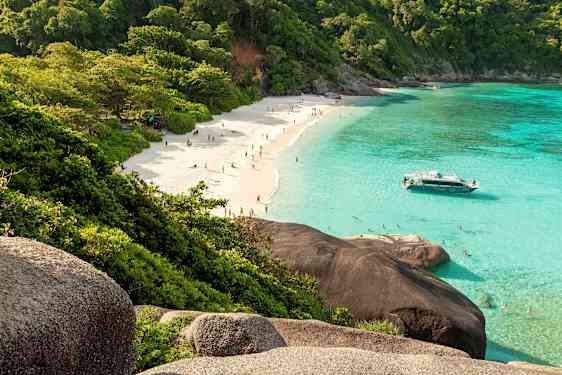 En sandstrand med människor, omgiven av grönskande träd och klart turkost vatten. En båt ligger nära stranden. (AI-genererad text)