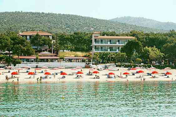 En strand med röda parasoller, solstolar och människor. Bakom stranden finns hotellbyggnader och skog. (AI-genererad text)