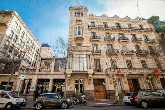 En gata med klassiska byggnader och parkerade bilar. Fönster och balkonger pryder fasaden. (AI-genererad text)