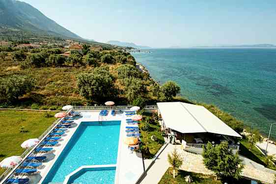 Utomhuspool med solsängar bredvid havet, omgivet av grönska och byggnader. (AI-genererad text)