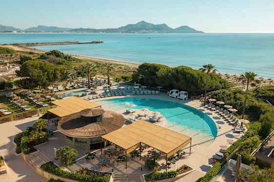 En swimmingpool omgiven av solstolar och parasoller, nära en strand och med utsikt över havet och berg i bakgrunden. (AI-genererad text)