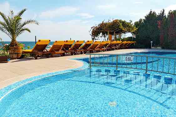 En swimmingpool med grunt vatten framför en rad gula solstolar på en terrass, med havet i bakgrunden. (AI-genererad text)