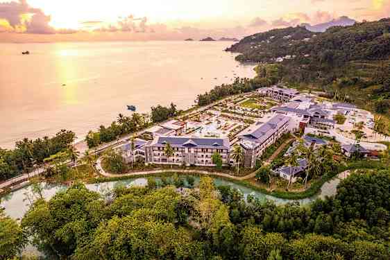 Flygbild över ett hotellkomplex omgivet av natur och beläget nära en kust med strand och hav vid solnedgång. (AI-genererad text)