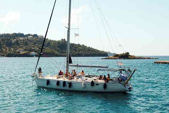 En segelbåt med flera personer ombord seglar nära en kustlinje med gröna kullar och några hus. (AI-genererad text)