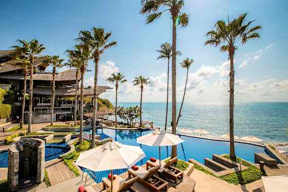 En poolområden vid havet med utsikt över klarblått vatten, omgivna av höga palmer. (AI-genererad text)