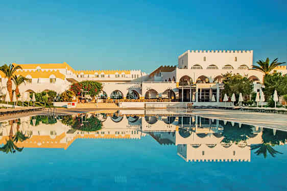 En vit byggnad med gult tak vid en pool med solstolar och parasoller. (AI-genererad text)