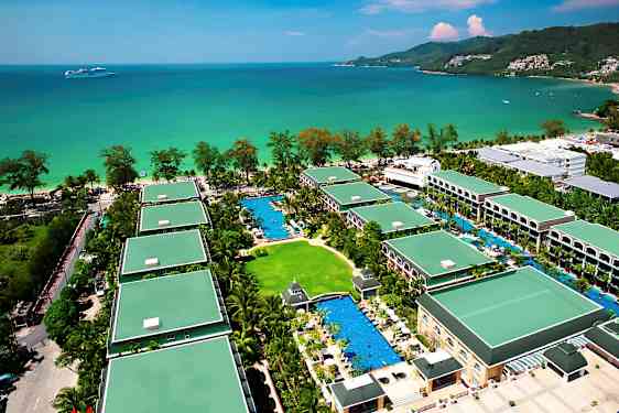 Flygvy över ett semesterhotell med flera byggnader omgivna av grönska och pooler, beläget vid havet. (AI-genererad text)