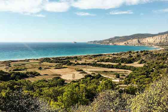 En landskapsbild med gröna fält, buskar och berg som sträcker sig mot en strand med turkost hav under en klar himmel. (AI-genererad text)