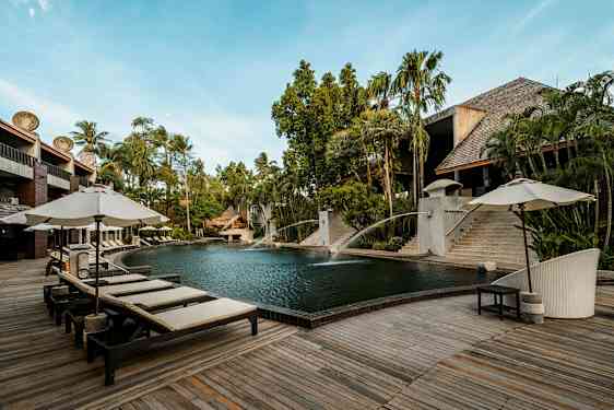 En utomhuspool omgiven av trädeck och solsängar med parasoller. Det finns frodig grönska och byggnader i trä och sten i bakgrunden. (AI-genererad text)