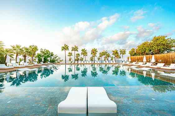 En utomhuspool med solstolar och parasoller, omgiven av palmer, och med speglingar i vattnet. (AI-genererad text)