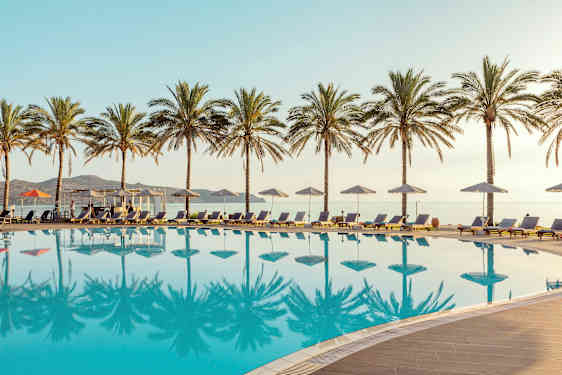 En pool med solstolar och parasoller, omgiven av palmer. Havet syns i bakgrunden. (AI-genererad text)