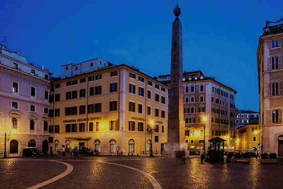 Ett torg med omgivande byggnader och en obelisk i mitten, upplyst på natten. (AI-genererad text)