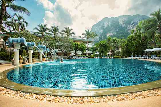 En simpool med klart blått vatten, omgiven av palmer och solstolar. I bakgrunden syns en klippa och himlen är delvis molnig. (AI-genererad text)