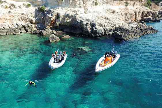Två båtar ligger för ankar i klart turkost vatten nära en klippig kust. Dykare är i vattnet. (AI-genererad text)